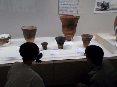 深鉢を横から観察する古代人と担当学芸員。